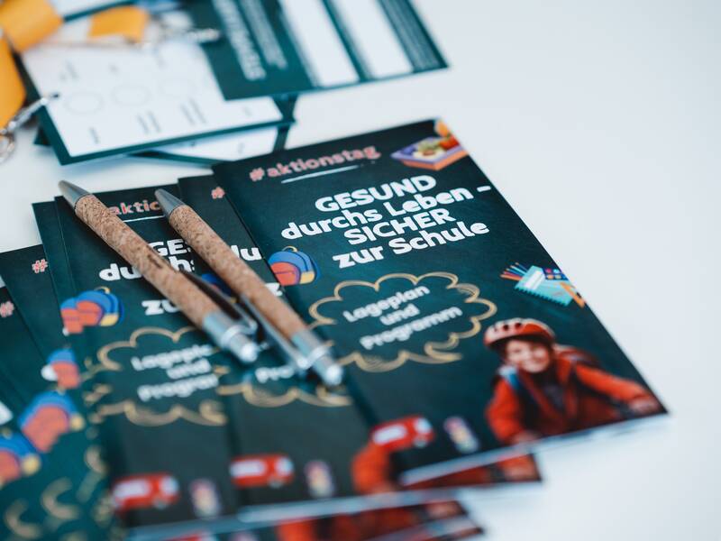 Ein Stapel von Broschüren mit dem Titel "GESUND durchs Leben - SICHER zur Schule" liegt auf einem Tisch, mit zwei Stiften darauf platziert.