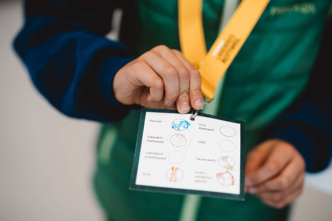 Eine Person hält ein laminiertes Blatt mit verschiedenen Logos und Kreisen in der Hand, das an einem gelben Umhängeband befestigt ist.