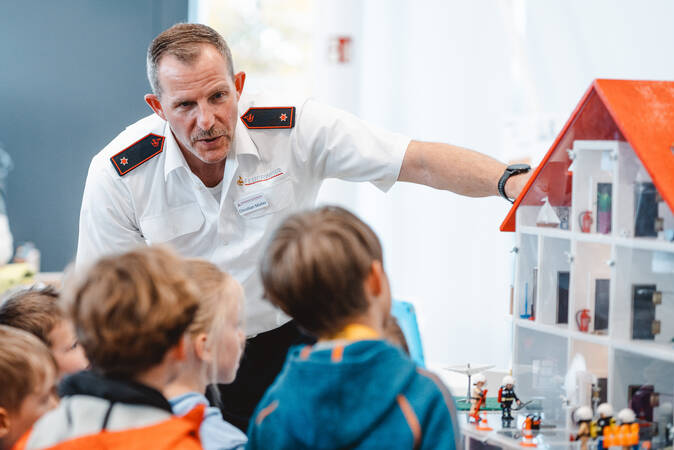 Ein Feuerwehrmann in Uniform erklärt Kinder spielerisch an einem großen Playmobil Haus wie man sich bei einem Hausbrand verhalten soll 