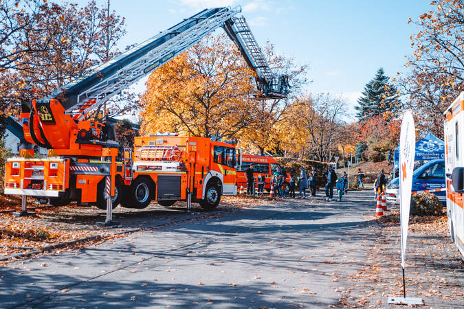 Eine fiktive Darstellung einer Einsatzübung mit mehreren Einsatzfahrzeugen der Feuerwehr und Menschen, die bei herbstlichem Wetter zuschauen.