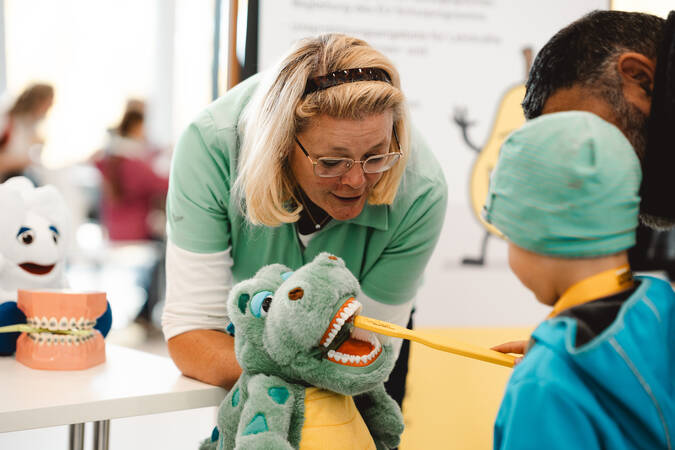 Eine Frau erklärt einem Kind und seinem Elternteil spielerisch an einem Krokoil wie man die Zähne richtig putzt