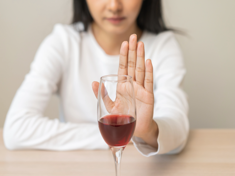 Eine Person in einem weißen Pullover hebt die Hand in einer ablehnenden Geste hinter einem Glas Rotwein.