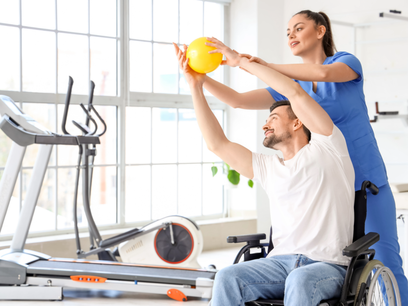 Ein Mann im Rollstuhl macht gemeinsam mit einer Physiotherapeutin eine Übung mit einem Ball