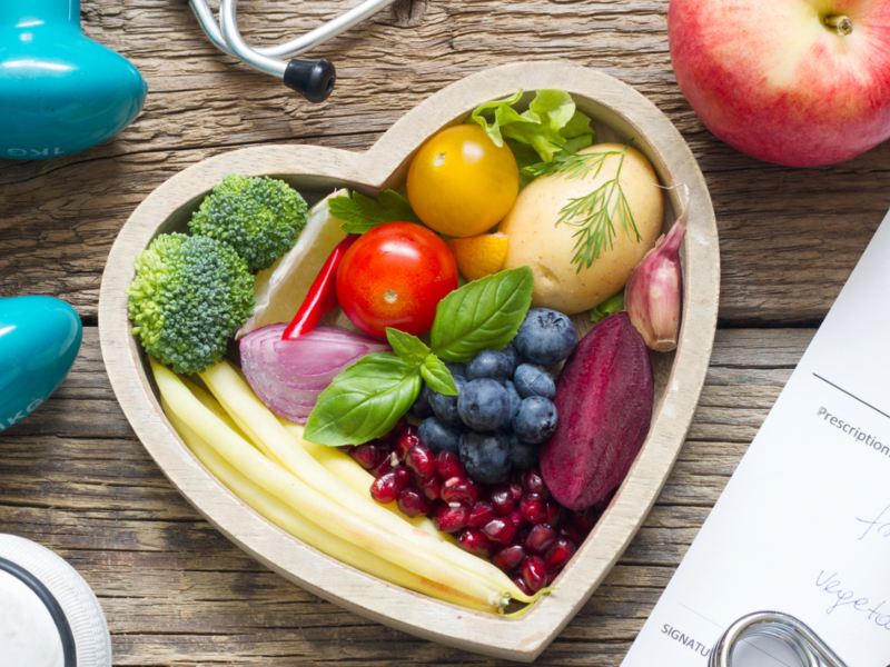 Ein Herz-förmiger Teller mit Obst und Gemüse, umgeben von Hanteln, einem Stethoskop, einem Apfel, einer Rezeptnotiz und Sportschuhen auf einem Holztisch.