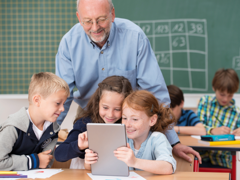 Kinder sind im Klassenzimmer und schauen gemeinsam interessiert auf ein Tablet und ein Lehrer steht hinter ihnen und schaut ebenfalls in das Tablett der Kinder