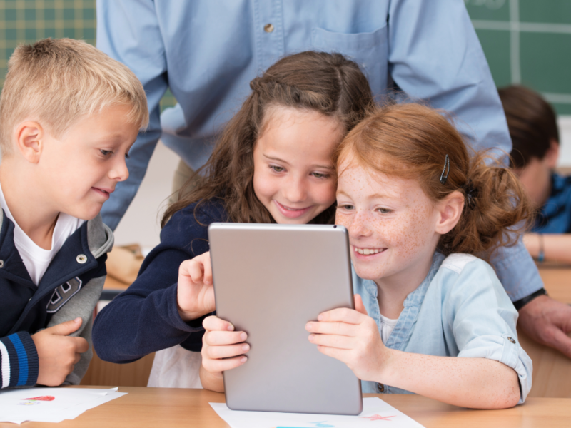 Kinder sind im Klassenzimmer und schauen gemeinsam interessiert auf ein Tablet und ein Lehrer steht hinter ihnen und schaut ebenfalls in das Tablett der Kinder